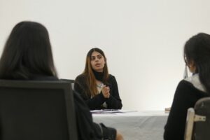A woman speaking to two colleagues across a small meeting table, delivering a good PowerPoint presentation without leaning on the slides